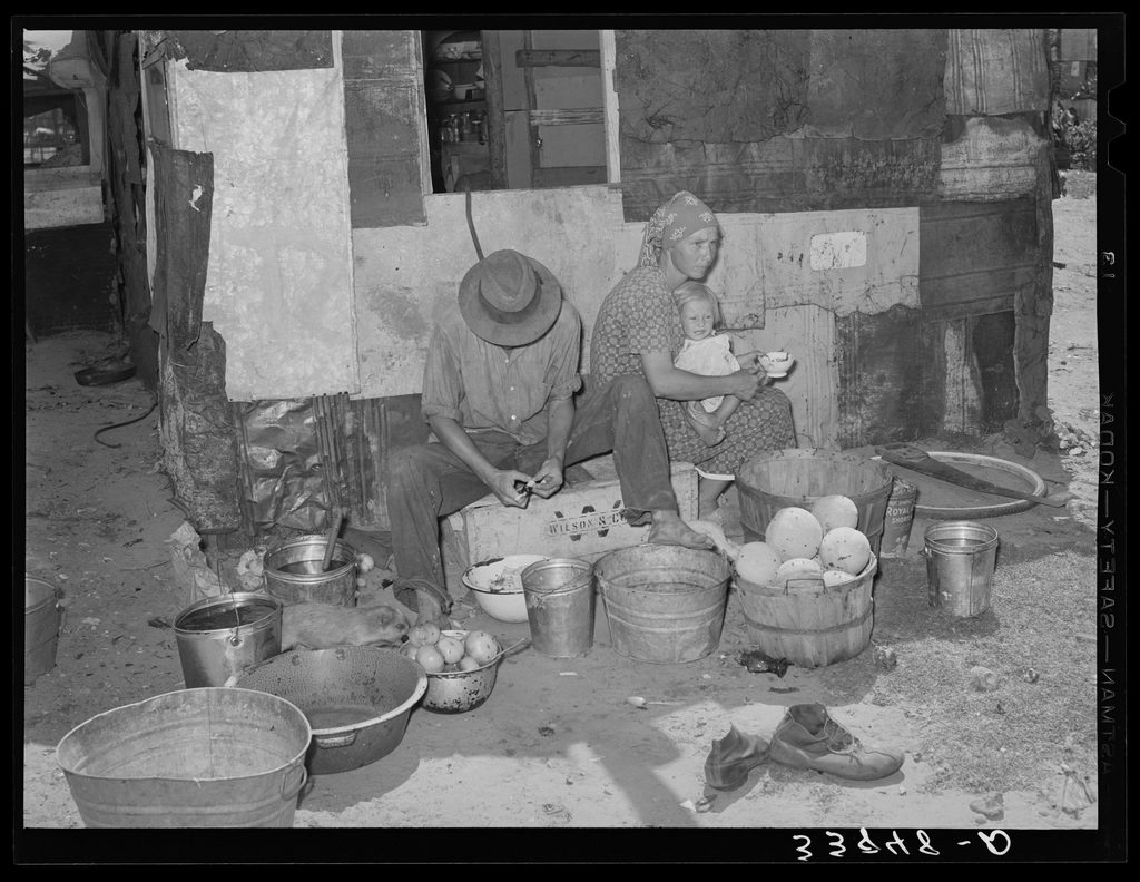 A family sits outside their makeshift shelter at a Depression-era camp, surrounded by scattered belongings.