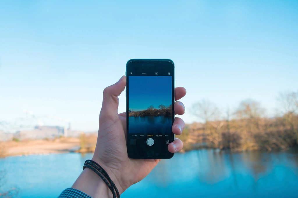 Person holding up a smartphone to photograph a calm lake, with hills and open sky in the distance